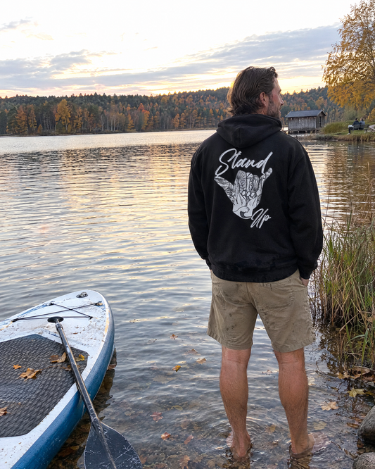 Stand Up Hang Loose Hoodie in Schwarz mit Rückenprint, getragen am Seeufer neben einem SUP-Board