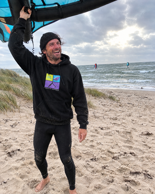 Wingfoil-Tools of Happiness Hoodie in Schwarz getragen am Ostseestrand, Wingfoiler hält den Wing über dem Kopf