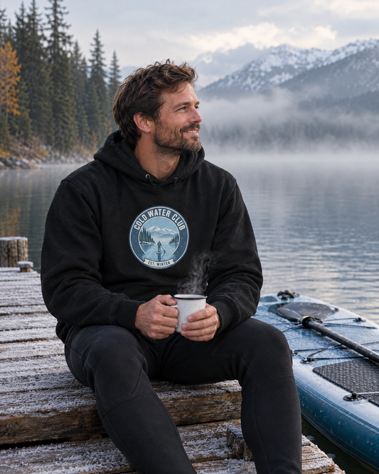 Mann im schwarzen Cold Water Club Hoodie sitzt mit dampfendem Becher auf einem frostigen Steg am nebligen Bergsee, daneben ein SUP-Board