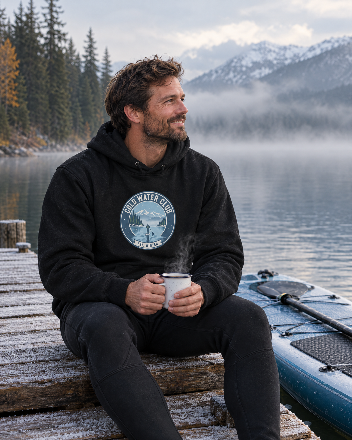 Mann im schwarzen Cold Water Club Hoodie sitzt mit dampfendem Becher auf einem frostigen Steg am nebligen Bergsee, daneben ein SUP-Board