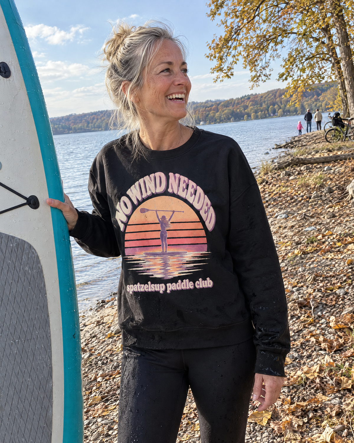 Frau im schwarzen No Wind Needed Pullover neben ihrem SUP-Board am See