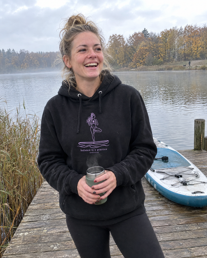 Frau im schwarzen Balance is a practice Hoodie am Steg nach dem SUP-Yoga