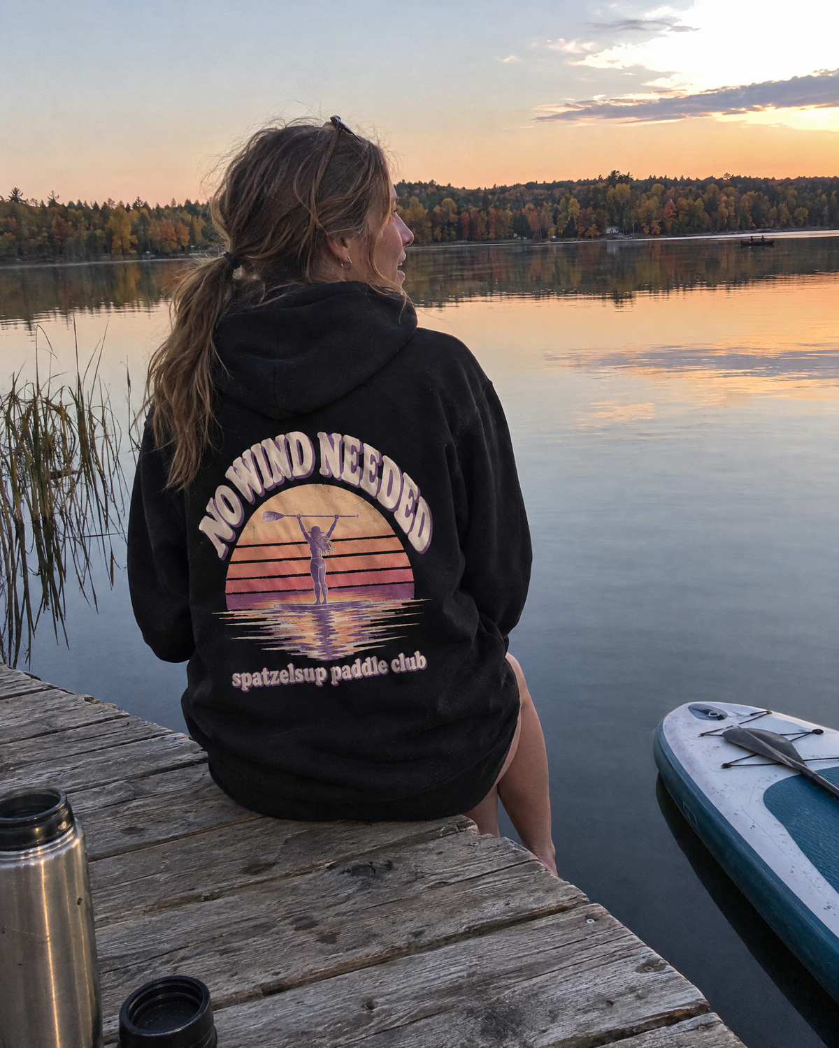 Frau im schwarzen No Wind Needed Hoodie sitzt am Steg, Rückenprint sichtbar