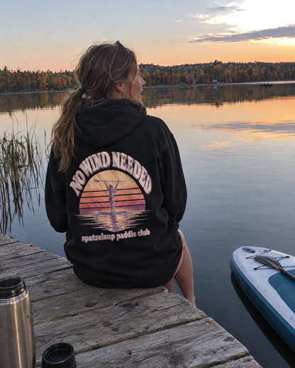 Frau im schwarzen No Wind Needed Hoodie sitzt am Steg, Rückenprint sichtbar